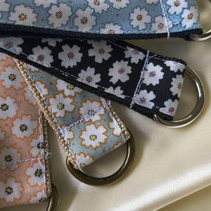Könnte beinhalten: Eine Sammlung von vier Stoffbändern mit einem sich wiederholenden weißen Blumenmuster mit gelben Zentren. Die Bänder sind in Hellblau, Schwarz, Pfirsich und Hellgrün gehalten und jeweils mit einem Metall-D-Ring versehen, der mit weißen Nähten befestigt ist.