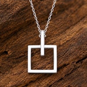 Könnte beinhalten: Eine silberfarbene Halskette mit einer feinen Kette und einem Anhänger in Form eines offenen Quadrats. Der Anhänger hängt an einem vertikalen Stab, der mit der Kette verbunden ist, und bildet ein geometrisches Design mit polierter Oberfläche.