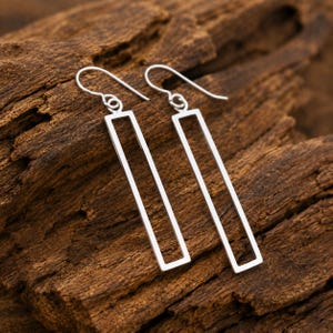 May include: A pair of silver-toned drop earrings featuring an open rectangular frame design. Each earring is suspended from a curved hook wire, creating a long, slender silhouette that hangs vertically from the top attachment point.