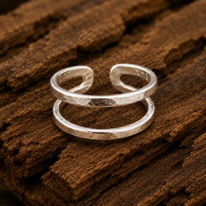 May include: A silver-toned open ring featuring a double-band design with a textured, hammered finish. The ring has a gap at the top, creating two parallel bands that connect at the sides, showcasing a metallic sheen and a rustic, uneven surface.