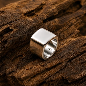 Op de afbeelding: Een gepolijste zilveren ring met een glad, vierkant oppervlak en een brede band. De binnenkant van de band is duidelijk gestempeld met het keurmerk 925, wat de metaalsamenstelling aangeeft. De ring heeft een reflecterende, metalen afwerking.