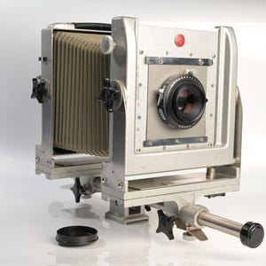 Op de afbeelding: Een grootformaat technische camera met een zilverkleurige metalen behuizing, een beige accordeonbalg en een zwarte lens op het voorpaneel. De camera heeft diverse zwarte instelknoppen, een rood rond logo en een metalen railbasis.