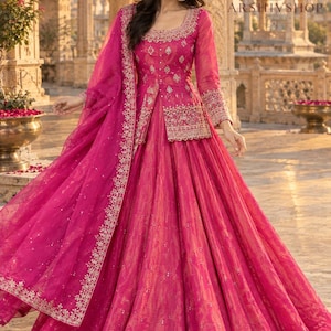 May include: A vibrant fuchsia floor-length gown featuring a long-sleeved bodice with intricate silver embroidery, a square neckline, and a matching sheer dupatta with a decorative border. The skirt is full and pleated with subtle metallic patterns.