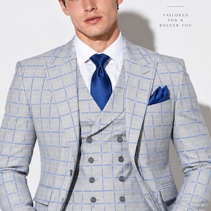 May include: A light gray three-piece suit with a blue windowpane check pattern, featuring a double-breasted waistcoat, a crisp white shirt, a solid blue necktie, and a matching blue pocket square. Text reads STITCH BY ARMAN TAILORED FOR A BOLDER YOU.
