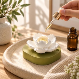 May include: A white ceramic lotus flower diffuser sits atop a smooth, olive green circular base. A glass dropper with a gold-toned collar releases a drop of liquid toward the center of the white petals, positioned near a small amber glass bottle.