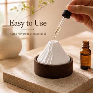May include: A white ceramic mountain-shaped essential oil diffuser sits inside a circular dark wood base. A glass dropper dispenses oil into the top opening. Text reads: Easy to Use. Add a few drops of essential oil. A brown glass bottle is nearby.