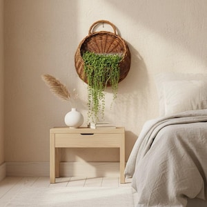 Può includere: Un cesto da parete rotondo in vimini intrecciato marrone contenente una pianta verde ricadente, posizionato sopra un comodino in legno chiaro con un vaso bianco contenente erba della pampa essiccata e libri, accanto a un letto con biancheria beige.