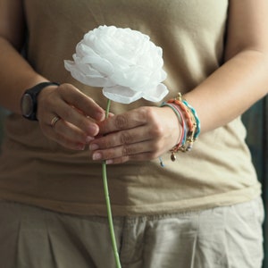 May include: A person holding a single white paper flower with a green stem. The person is wearing a light brown tank top and several bracelets on their wrist.