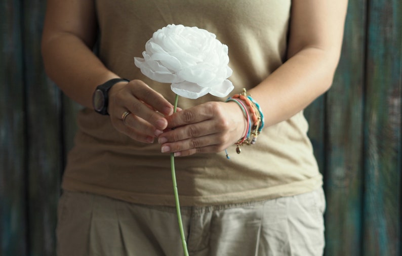 May include: A person holding a single white paper flower with a green stem. The person is wearing a light brown tank top and several bracelets on their wrist.