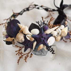Può includere: Un cerchietto nero con una base in rete nera decorata con conchiglie, stelle marine e fogliame secco. Le conchiglie sono bianche, marroni e viola.