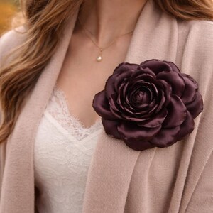 May include: A large, dark burgundy satin rose brooch pinned to a pale pink cardigan. The brooch is the focal point, with detailed petals. A white lace top and gold necklace with a pearl pendant are also visible.