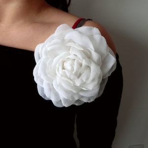 May include: A large white fabric flower with multiple layers of petals is pinned to a black off-the-shoulder top.