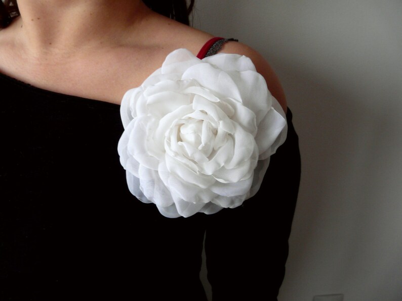 May include: A large white fabric flower with multiple layers of petals is pinned to a black off-the-shoulder top.