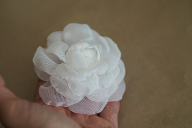 May include: A white fabric flower with a delicate, layered design. The flower is made of soft, sheer fabric and has a slightly ruffled appearance.
