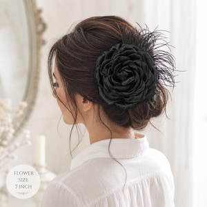 Könnte beinhalten: Ein schwarzer Satin-Blumen-Haarclip mit Federakzenten. Die Blume hat einen Durchmesser von 18 cm und wird im Haar getragen. Die Frau trägt ein weißes Hemd mit Knöpfen.