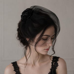 Könnte beinhalten: Ein schwarzer Fascinator-Kopfschmuck mit einem strukturierten Netzschleier, der das Gesicht teilweise bedeckt, und einer dekorativen Stoffrose an der Seite. Das Accessoire besteht aus feinem schwarzem Netzstoff und einem passenden Blumenelement.