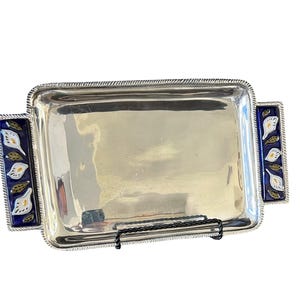 May include: A rectangular silver-toned serving tray with a polished, reflective surface and a rope-textured border. The tray features two side handles decorated with deep blue ceramic inlays, each displaying a pattern of white calla lilies.