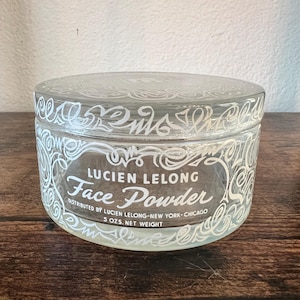 May include: A clear glass jar with a lid featuring white decorative scrollwork patterns. The front text reads LUCIEN LELONG Face Powder, DISTRIBUTED BY LUCIEN LELONG-NEW YORK-CHICAGO, and 5 OZS. NET WEIGHT. The container has a cylindrical shape.