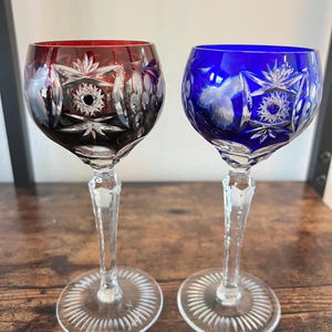May include: Two cut-to-clear crystal wine glasses, one in deep red and one in vibrant blue. Each glass features intricate starburst and leaf patterns etched into the colored glass, supported by a clear, faceted stem and a circular base.