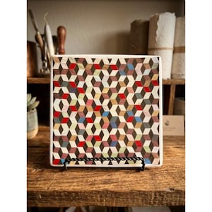 May include: A square ceramic tile featuring a repeating geometric pattern of small diamonds in shades of brown, white, red, and blue, creating a three-dimensional cube effect. The tile is displayed upright on a black metal wire stand.