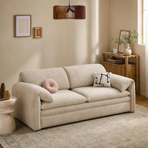 May include: A beige upholstered sofa with two large seat cushions and two back cushions. The sofa features rounded armrests and includes one round, tufted pink throw pillow and one rectangular white throw pillow with a black abstract line design.