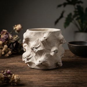May include: A cylindrical off-white ceramic vessel featuring a textured surface adorned with multiple sculpted skull motifs integrated into swirling, raised patterns that wrap around the entire exterior of the container.