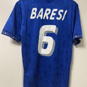 Könnte beinhalten: Ein blaues Fußballtrikot mit dem Namen BARESI und der Nummer 6 in Weiß auf der Rückseite. Das Trikot hat ein dezentes Ton-in-Ton-Muster im gesamten Stoff, einen Kragen und Ärmelbündchen mit kleinen roten, weißen und grünen Dreiecksdetails.