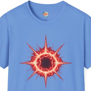 May include: A light blue short-sleeve t-shirt featuring a central graphic of a glowing red and orange starburst or sun-like shape with sharp, radiating points and a dark, circular center. The design has a vibrant, energetic appearance.
