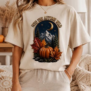 Peut inclure: Un t-shirt beige à manches courtes avec une impression graphique représentant une citrouille, des feuilles d'automne, des pins et un croissant de lune sous un ciel nocturne. Le texte "INTO THE AUTUMN WILD" est arqué au-dessus de l'illustration.