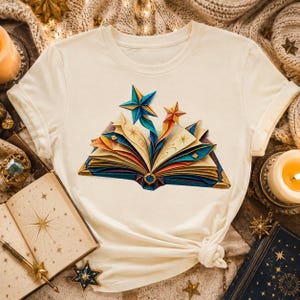 Peut inclure: Un t-shirt couleur crème arborant un motif de livre ouvert dont les pages se transforment en étoiles de style origami bleues et orange. Le design présente des lignes dorées complexes et des motifs d'étoiles sur les pages, centré sur la poitrine.