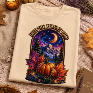 Peut inclure: Un t-shirt beige avec une impression graphique représentant une citrouille, des feuilles d'automne colorées et un paysage de montagne sous un croissant de lune et un ciel étoilé. Le texte "INTO THE AUTUMN WILD" est arqué au-dessus.