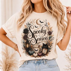 Peut inclure: Un t-shirt couleur crème arborant une impression graphique avec le texte Spooky Season en écriture noire. Le motif comprend un croissant de lune, des chauves-souris, des roses noires, une lanterne, une tasse avec chantilly, un chat noir et des citrouilles.