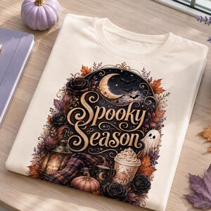 Peut inclure: Un t-shirt couleur crème avec une impression graphique portant le texte Spooky Season dans une police décorative. Le motif comprend un croissant de lune, des chauves-souris, un fantôme, une lanterne, une tasse de café, des citrouilles et des fleurs.