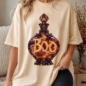 Peut inclure: Un t-shirt beige à manches courtes orné d'un motif central représentant une bouteille de potion en verre sombre et ornementée. À l'intérieur, une fumée orange et jaune tourbillonnante forme le mot "BOO" avec un effet ardent et un bouchon en cristal.