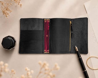 May include: A black leather notebook cover featuring a central spine with bright pink elastic cords held by gold-toned eyelets. The interior includes a vertical zippered pocket with a gold zipper pull, a pen loop, and two horizontal storage slots.