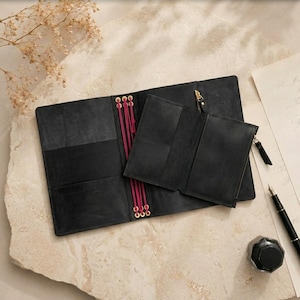 May include: A black leather notebook cover featuring a central spine with bright pink elastic cords and brass eyelets. A matching black leather zippered pouch is attached to the cover, which includes internal card slots and a gold-toned zipper pull.