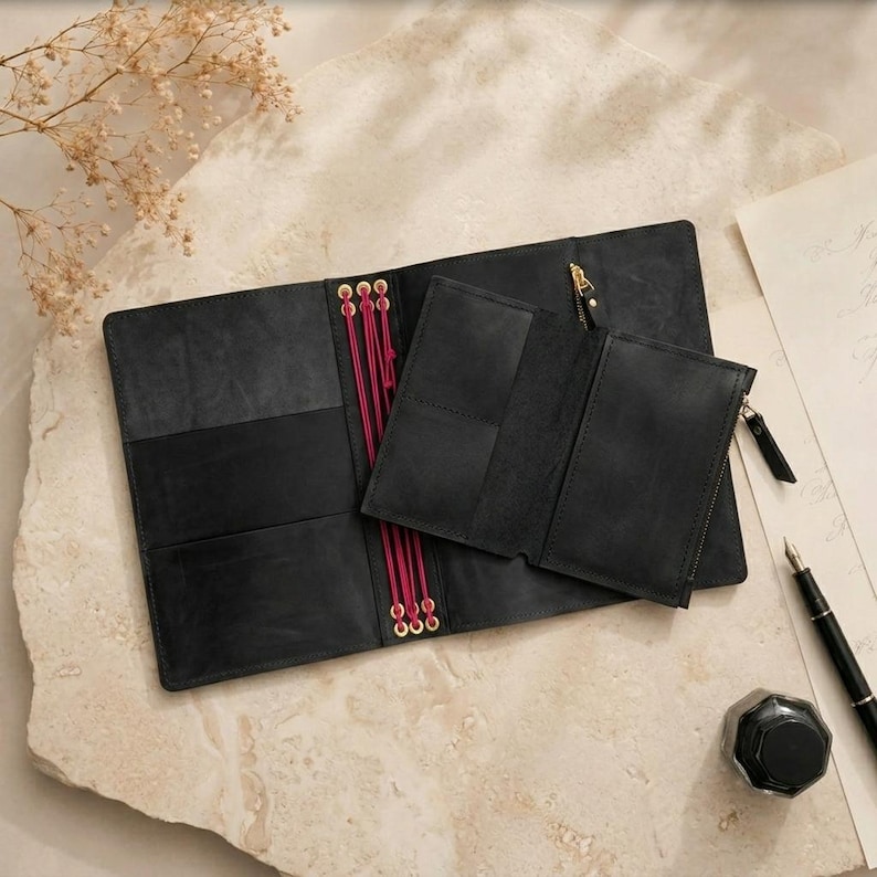 May include: A black leather notebook cover featuring a central spine with bright pink elastic cords and brass eyelets. A matching black leather zippered pouch is attached to the cover, which includes internal card slots and a gold-toned zipper pull.