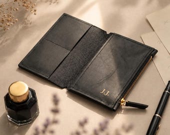 May include: A black leather wallet opened to reveal interior card slots and a zippered compartment. The exterior features the gold embossed initials J.J. and a gold metal zipper pull. The wallet is displayed alongside a black ink bottle and pen.