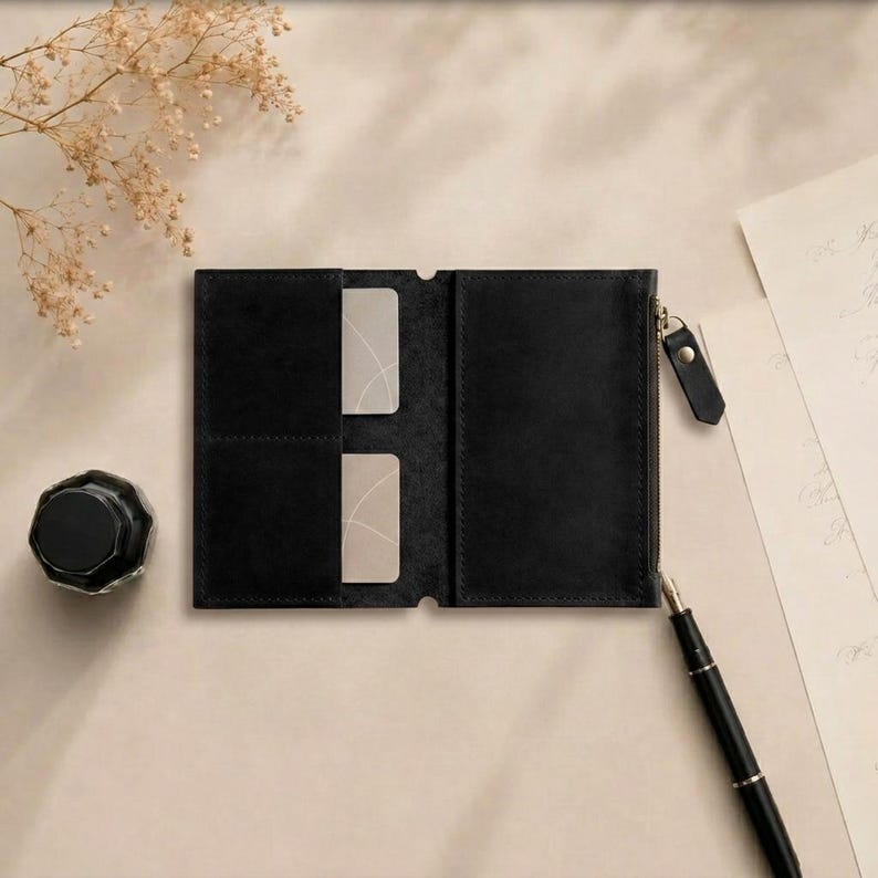 May include: A black leather wallet featuring two interior card slots and a side zipper compartment with a black pull tab. The wallet is open, revealing a smooth matte texture and clean stitched edges, designed for holding cards and small items.