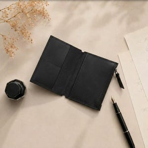 May include: A black leather wallet featuring a bi-fold design with multiple interior card slots and a side zipper closure. The wallet is positioned flat, showcasing its smooth texture, stitched edges, and a functional metal zipper pull.