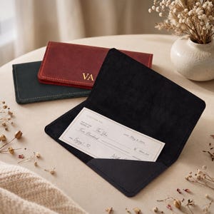 May include: Three rectangular leather checkbook covers in black and burgundy. One black cover is open, displaying a gift check with the text A GIFT FOR YOU, PAY TO THE ORDER OF, DATE May 9, 2024, and $ 500.00. The burgundy cover features the gold initials VA.