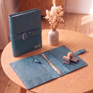 May include: A teal leather binder with a buckle strap closure and the gold embossed initials B.M. The binder is shown both closed and open, revealing a three-ring metal mechanism, interior pockets, a wooden handle stamp, and an ink pad.