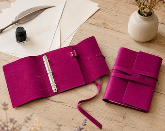 May include: Two magenta leather journals, one open and one closed. The open journal features a three-ring metal binder mechanism and a leather strap closure. The closed journal is secured with a matching wrap-around leather tie.
