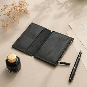 May include: A black leather wallet with a bi-fold design, featuring an open interior with card slots on the left side and a zippered compartment on the right side. The wallet has a smooth texture and is positioned next to a fountain pen and ink.