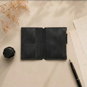 May include: A black leather wallet opened flat, featuring a rectangular pocket on the left side and a zippered compartment on the right side. The wallet has a smooth, matte texture and is positioned alongside a black inkwell and a fountain pen.
