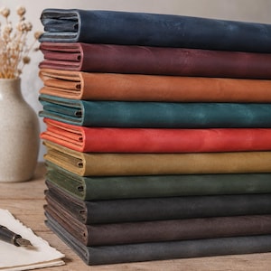Könnte beinhalten: Ein vertikaler Stapel aus zehn rechteckigen Lederjournalen oder Notizbüchern in verschiedenen Farben, darunter Marineblau, Burgunderrot, Hellbraun, Blaugrün, Rot, Senfgelb, Olivgrün, Anthrazit, Dunkelbraun und Schwarz mit einer matten Oberfläche.