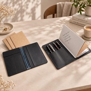 May include: Two black leather notebook covers with internal pockets and elastic bands. One cover is open, holding a lined notebook with handwritten text and three pens in dedicated slots. The other cover is empty, showing the interior layout.