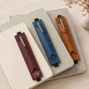 Könnte beinhalten: Drei rechteckige Stiftehalter aus Leder in Burgunderrot, Blau und Hellbraun, jeweils mit einem silbernen Metallstift und einem elastischen Band. Der burgunderrote Halter ist mit SW, der blaue mit JB und der hellbraune mit Rick geprägt.