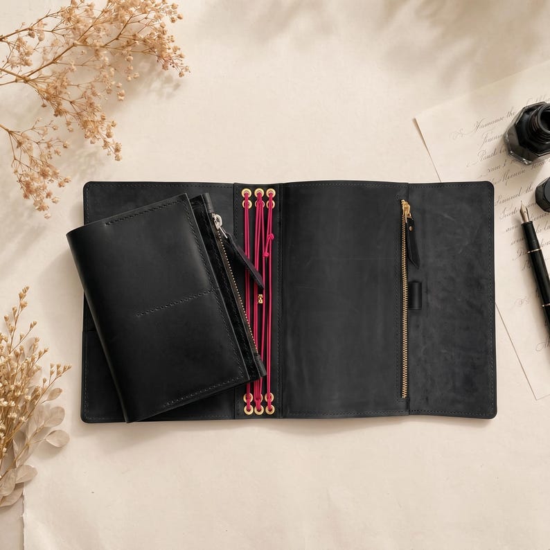 May include: A black leather traveler's notebook cover featuring a zippered pocket on the left panel, a vertical zippered compartment on the right panel, and bright pink elastic cords threaded through gold-toned eyelets at the center spine.