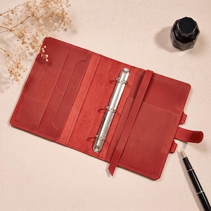 May include: An open red leather binder featuring a silver three-ring metal mechanism in the center. The interior includes multiple card slots on the left side, a vertical strap on the right, and a secure tab closure on the outer edge of the cover.